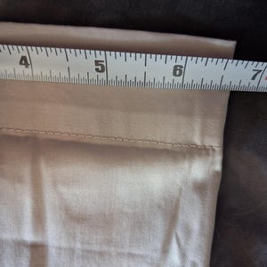 May include: A beige fabric item is measured with a white and black measuring tape. The tape shows a measurement of approximately 7 inches. The fabric has a stitched seam near the top edge.