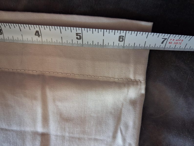 May include: A beige fabric item is measured with a white and black measuring tape. The tape shows a measurement of approximately 7 inches. The fabric has a stitched seam near the top edge.