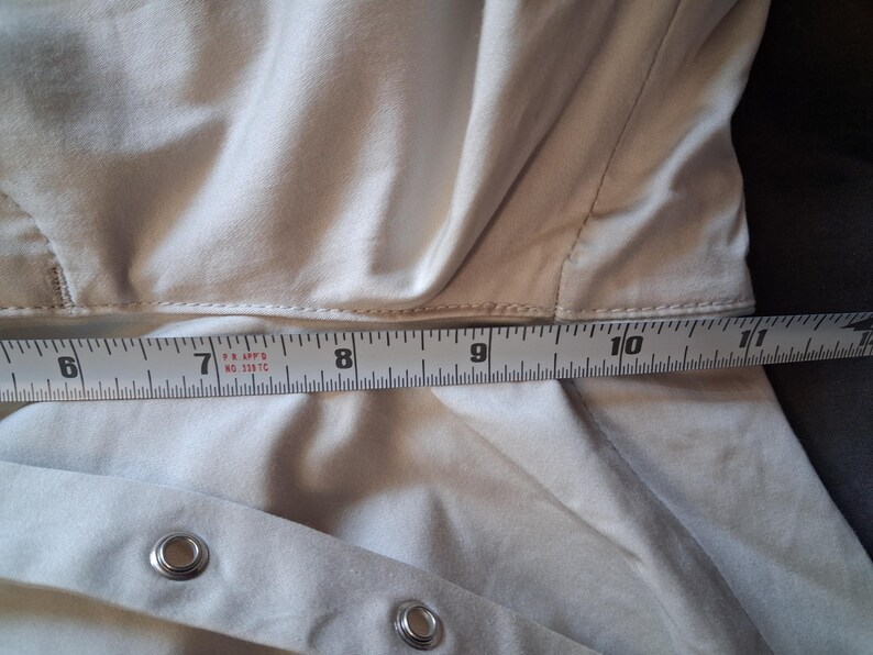May include: Close-up of a beige garment with a measuring tape laid across it. The tape shows measurements from 6 to 11 inches. The garment has metal eyelets and a seam running across the middle.
