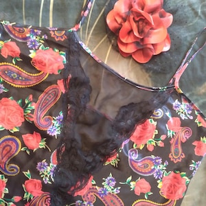 May include: A black satin camisole with a floral and paisley pattern in red, purple, and yellow. The camisole has a black lace trim and spaghetti straps. A large red fabric flower is also visible.