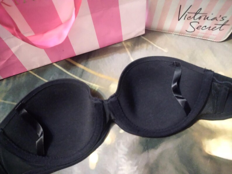 May include: A black strapless bra with a smooth texture. The bra has a classic design with molded cups and a band for support. A pink and white striped shopping bag and a small bag with the text "Victoria's Secret" are in the background.