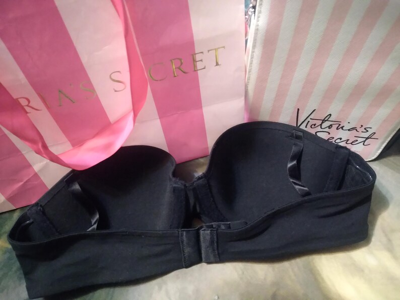 May include: A black strapless bra with a clasp closure. The bra is displayed in front of two pink and white striped shopping bags with the words "Victoria's Secret" printed on them.