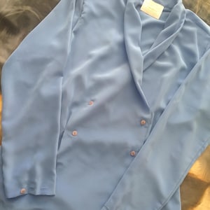 May include: A light blue blazer with a notched lapel, long sleeves, and button closures. The blazer has a double-breasted design with four buttons and a small tag near the collar. The fabric appears to be a smooth, lightweight material.