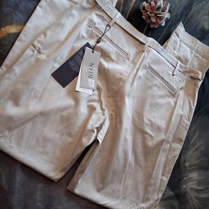 May include: A pair of beige-colored pants with a tag attached. The pants have a classic design with a belt and pockets. The tag reads "NYDJ". A decorative flower is also present.