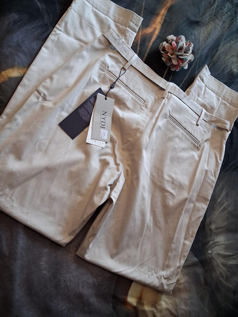 May include: A pair of beige-colored pants with a tag attached. The pants have a classic design with a belt and pockets. The tag reads "NYDJ". A decorative flower is also present.