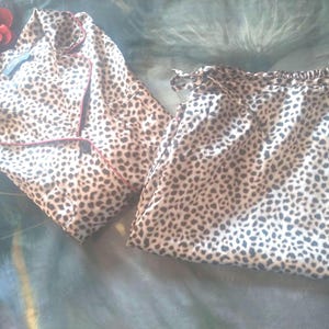 May include: A two-piece pajama set with a leopard print pattern. The top is a button-down shirt with red trim, and the bottoms are drawstring pants. A red flower is in the upper left corner. The pajamas are made of a soft, silky material.