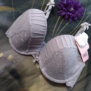 May include: A light gray bra with lace detailing and a bow on each strap. The bra has a soft, gathered fabric and a delicate lace trim. A purple flower is next to the bra.