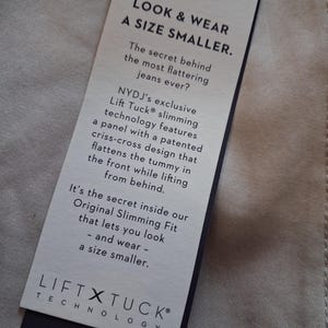 May include: A rectangular tag with the text "LOOK & WEAR A SIZE SMALLER" in black font. The tag also includes the text "LIFT X TUCK TECHNOLOGY" and "NYDJ PREMIUM DENIM". The tag is attached to a beige fabric.