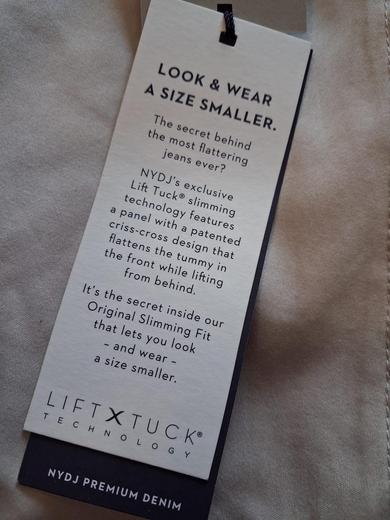 May include: A rectangular tag with the text "LOOK & WEAR A SIZE SMALLER" in black font. The tag also includes the text "LIFT X TUCK TECHNOLOGY" and "NYDJ PREMIUM DENIM". The tag is attached to a beige fabric.