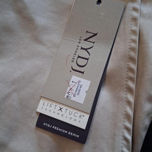 May include: Close-up of tan-colored pants with a tag attached. The tag reads "NYDJ LOS ANGELES" and includes the text "LIFT X TUCK TECHNOLOGY" and "NYDJ PREMIUM DENIM". The pants are folded and have visible stitching.
