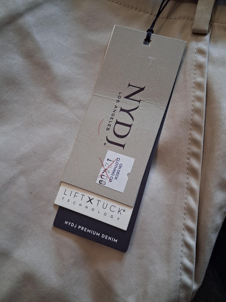 May include: Close-up of tan-colored pants with a tag attached. The tag reads "NYDJ LOS ANGELES" and includes the text "LIFT X TUCK TECHNOLOGY" and "NYDJ PREMIUM DENIM". The pants are folded and have visible stitching.