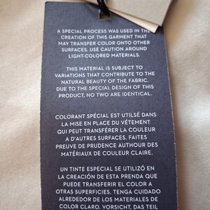 May include: A black tag with text in English, French, Spanish, and German, warning about potential color transfer from the garment. The tag is attached to a beige fabric with a black string.