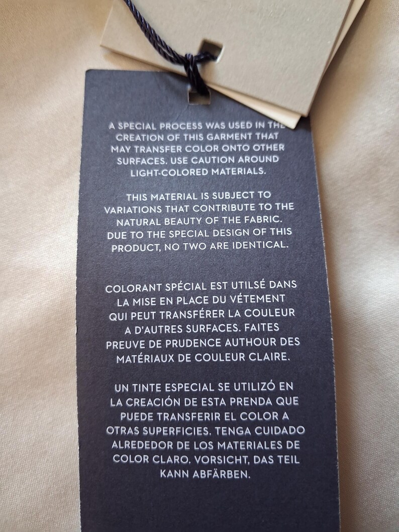 May include: A black tag with text in English, French, Spanish, and German, warning about potential color transfer from the garment. The tag is attached to a beige fabric with a black string.