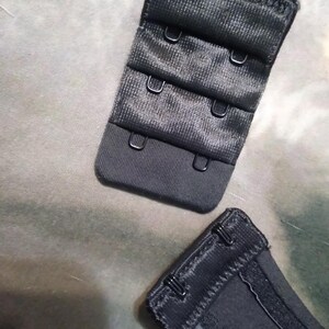 May include: Close-up of black bra hook and eye closures. The closures are made of a textured fabric and feature multiple hook and eye settings for adjustable sizing. The metal hooks are silver.