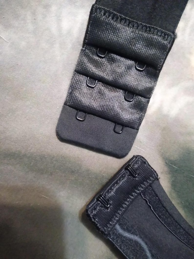 May include: Close-up of black bra hook and eye closures. The closures are made of a textured fabric and feature multiple hook and eye settings for adjustable sizing. The metal hooks are silver.