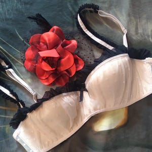 May include: Ivory-colored bra with black lace trim and straps, adorned with small white polka dots. A large red fabric flower with black accents rests nearby. The bra features a bow detail and a delicate, feminine design.