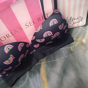 May include: Black bra with a rainbow pattern in pink, yellow, and blue. The bra has adjustable straps and a seamless design. A pink and white striped shopping bag is in the background.