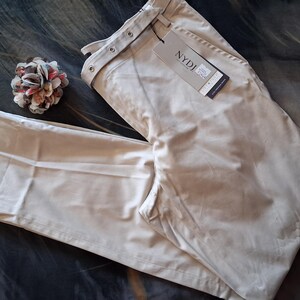 May include: Beige-colored pants with a tag that reads "NYDJ". The pants are folded and lying on a patterned surface. A decorative flower pin is also visible.