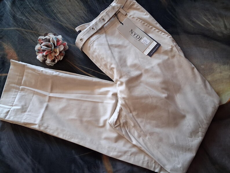 May include: Beige-colored pants with a tag that reads "NYDJ". The pants are folded and lying on a patterned surface. A decorative flower pin is also visible.