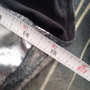 May include: A close-up of a silver fabric with a black seam. A white measuring tape is laid across the fabric, showing measurements from 14 to 16 inches.