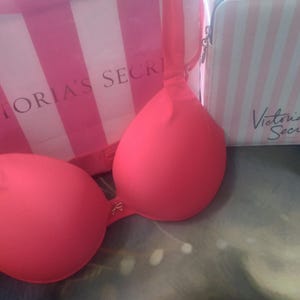 May include: A bright coral-colored bra with a matching pink Victoria's Secret shopping bag and a small striped cosmetic bag. The bra has a delicate clasp and adjustable straps. The shopping bag and cosmetic bag have the Victoria's Secret logo.