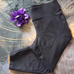 May include: Black leggings with a wide waistband. A purple flower with dark accents is placed near the leggings. The leggings are made of a smooth, opaque fabric. The leggings are folded.
