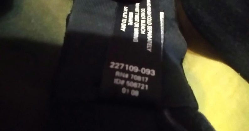 May include: A black clothing tag with white text. The tag includes care instructions: "HAND WASH COLD SEPARATELY, DO NOT BLEACH, DO NOT TWIST OR WRING, RESHAPE, LAY FLAT TO DRY." It also has the numbers "227109-093", "RN# 70817", "ID# 506721", and "01 08".