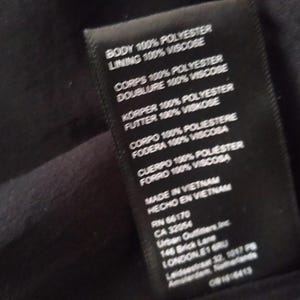 May include: Black clothing label with white text detailing the fabric composition and origin of the garment. The label reads "Body 100% Polyester, Lining 100% Viscose, Corps 100% Polyester, Doublure 100% Viscose, K&ouml;rper 100% Polyester, Futter 100% Viskose, Corpo 100% Poliestere, Fodera 100% Viscosa, Cuerpo 100% Poli&eacute;ster, Forro 100% Viscosa, Made in Vietnam, Hecho en Vietnam, RN 66170, CA 32054, Urban Outfitters, Inc., 146 Brick Lane, London, E1 6RU, Leidsestraat 32, 1017 PB, Amsterdam, Netherlands, 0814-6413".