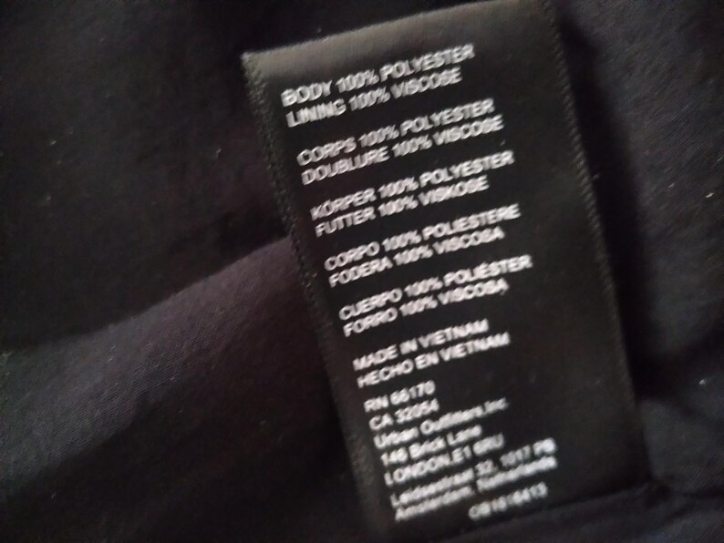 May include: Black clothing label with white text detailing the fabric composition and origin of the garment. The label reads "Body 100% Polyester, Lining 100% Viscose, Corps 100% Polyester, Doublure 100% Viscose, K&ouml;rper 100% Polyester, Futter 100% Viskose, Corpo 100% Poliestere, Fodera 100% Viscosa, Cuerpo 100% Poli&eacute;ster, Forro 100% Viscosa, Made in Vietnam, Hecho en Vietnam, RN 66170, CA 32054, Urban Outfitters, Inc., 146 Brick Lane, London, E1 6RU, Leidsestraat 32, 1017 PB, Amsterdam, Netherlands, 0814-6413".