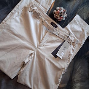 May include: Beige-colored pants with a belt featuring metal eyelets and a buckle. The pants have a tag that reads "NYDJ". A decorative flower is placed near the pants.