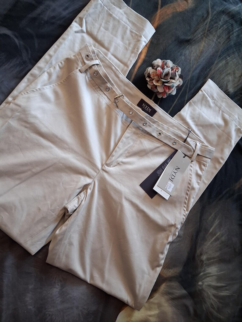 May include: Beige-colored pants with a belt featuring metal eyelets and a buckle. The pants have a tag that reads "NYDJ". A decorative flower is placed near the pants.