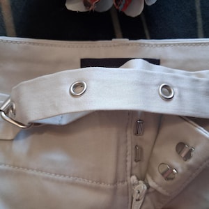 May include: Close-up of light beige pants with a matching belt. The belt has silver grommets and a silver buckle. The pants have a zipper and two silver button closures.