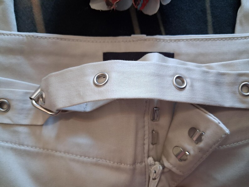 May include: Close-up of light beige pants with a matching belt. The belt has silver grommets and a silver buckle. The pants have a zipper and two silver button closures.