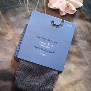 May include: A pair of dark gray textured tights in original packaging. The blue cardboard header features the text SimplyVera Vera Wang and Tights, with Size 1 and Made in USA printed in the corner. A pale pink fabric flower sits above.