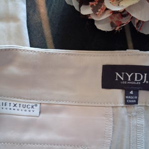 May include: Close-up of a pair of light beige pants. The pants have a tag that reads "NYDJ LOS ANGELES" and "LIFT X TUCK TECHNOLOGY". The size is 4, and the pants are made in China. The fabric appears to be a smooth, woven material.