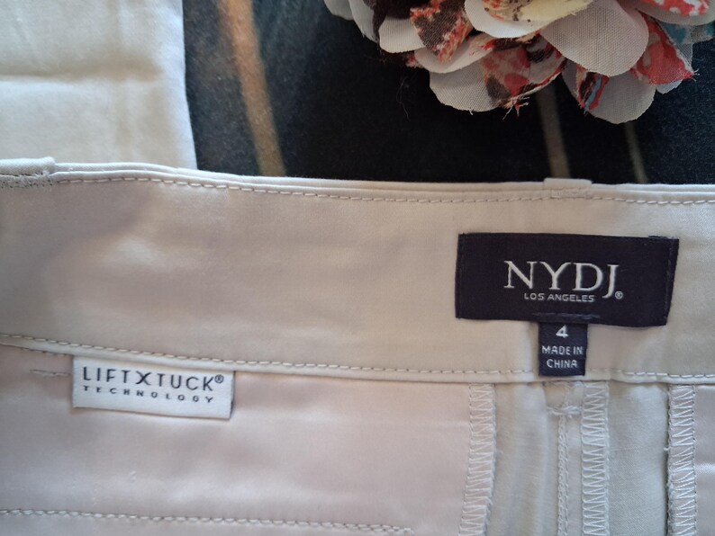 May include: Close-up of a pair of light beige pants. The pants have a tag that reads "NYDJ LOS ANGELES" and "LIFT X TUCK TECHNOLOGY". The size is 4, and the pants are made in China. The fabric appears to be a smooth, woven material.