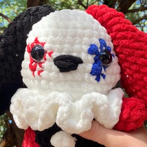 May include: A crocheted plush toy with a white body, black and red ears, and black and blue eyes. The toy has a black nose and a red and blue design on its face.