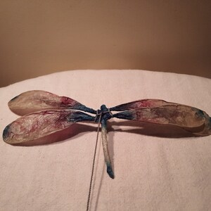 May include: A handcrafted dragonfly sculpture with delicate wings. The wings are a translucent beige with red and blue accents. The body is a light beige with a blue tail. A thin wire extends from the body.