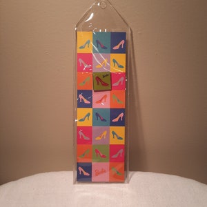 May include: A colorful Barbie-themed notepad featuring a grid pattern of high-heeled shoe silhouettes in various colors. The notepad is encased in a clear plastic sleeve with a small Barbie logo on the front.