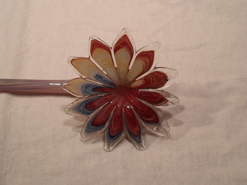 May include: A glass flower-shaped object with a long stem. The flower petals are a mix of red, yellow, and blue, with a clear glass outline. The stem is a pale purple colour.