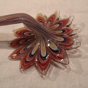 May include: A decorative glass flower with a long, curved stem. The flower petals are a mix of red, yellow, blue, and white, with a clear glass edge. The stem has a similar colour pattern.