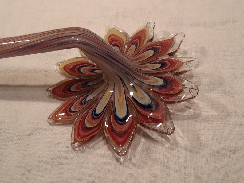 May include: A decorative glass flower with a long, curved stem. The flower petals are a mix of red, yellow, blue, and white, with a clear glass edge. The stem has a similar colour pattern.
