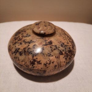 May include: A speckled, rounded stone container with a small, circular lid. The container is primarily tan with dark brown and black spots. The lid has a carved design on top. The container sits on a white surface.