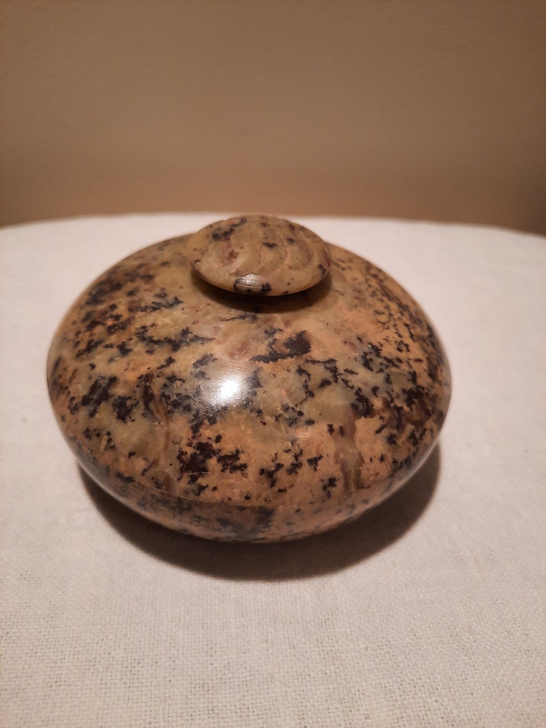 May include: A speckled, rounded stone container with a small, circular lid. The container is primarily tan with dark brown and black spots. The lid has a carved design on top. The container sits on a white surface.