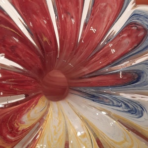 May include: A decorative glass bowl with a flower-like pattern. The bowl has red, white, yellow, and blue swirls. The centre of the bowl has a small, round, reddish-brown glass ball.