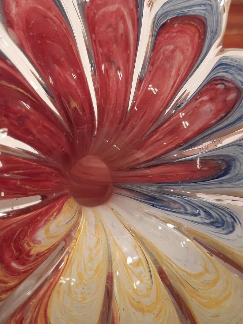 May include: A decorative glass bowl with a flower-like pattern. The bowl has red, white, yellow, and blue swirls. The centre of the bowl has a small, round, reddish-brown glass ball.
