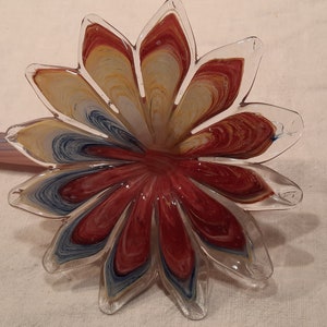 May include: A clear glass flower-shaped ornament with red, yellow, and blue swirls. The petals are transparent, and the centre is a deep red. A glass stem is visible in the background.