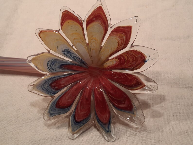 May include: A clear glass flower-shaped ornament with red, yellow, and blue swirls. The petals are transparent, and the centre is a deep red. A glass stem is visible in the background.