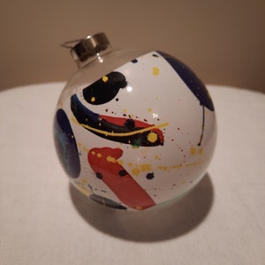 May include: A clear glass Christmas ornament with a silver metal cap. The ornament is white with colorful abstract paint splatters in red, yellow, blue, and black. The ornament is round and has a shiny finish.