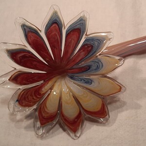 May include: A clear glass flower-shaped object with red, yellow, and blue swirls. The flower has a long, thin stem with pink and blue stripes. The petals are detailed with colour variations.